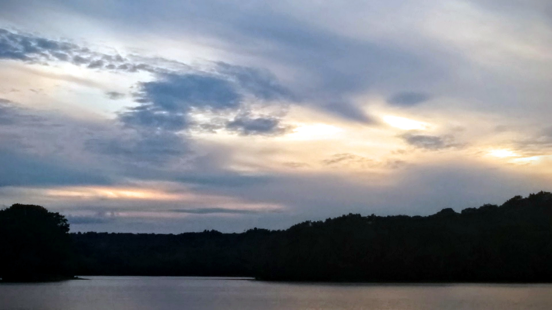 Wide-angle waterscape at dusk of Spy Pond, in Arlington, Massachusetts, USA. The crepuscular sky occupies most of the width and height of the photo, with thin white and dark clouds. Across the bottom is the water and the silhouetted opposite bank of the pond.